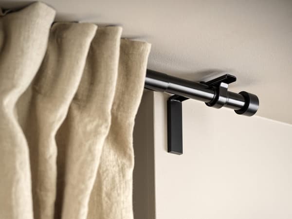 A black curtain rod with cream-colored curtains pleated and hanging on it.