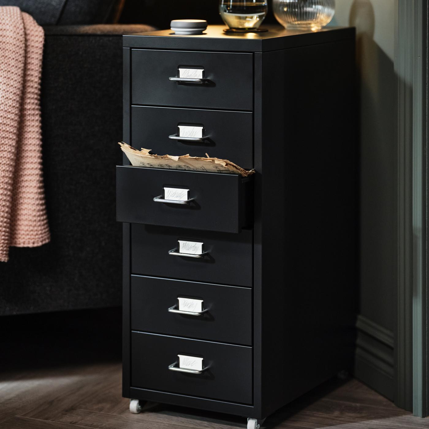 A black HELMER drawer unit on castors at an angle between a wall and a sofa, a pale-pink INGABRITTA throw over the armrest.