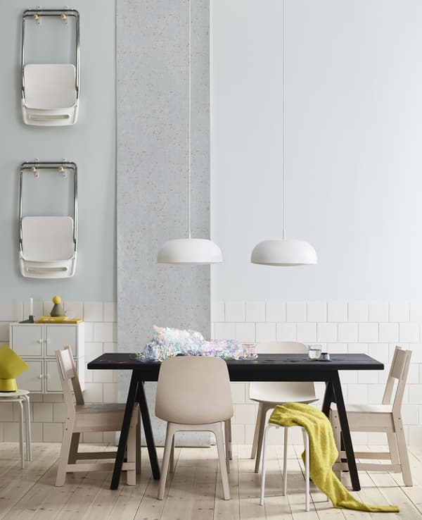 A black IKEA dining table with six different white and beige seats and two white pendant lamps hanging overhead.