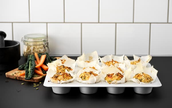 A black kitchen worktop with a VARDAGEN metal muffin tin filled with twelve muffins made with carrots and pumpkin seeds.
