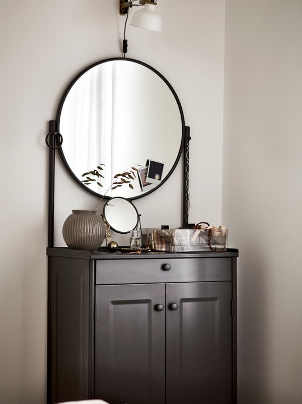 A black KORNSJÖ cabinet with a large, round mirror, displaying decorative objects and a white lamp placed above it.