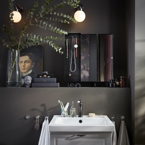 A black mirror, a vase with eucalyptus, two round wall-mounted lamps and a white wash-basin.