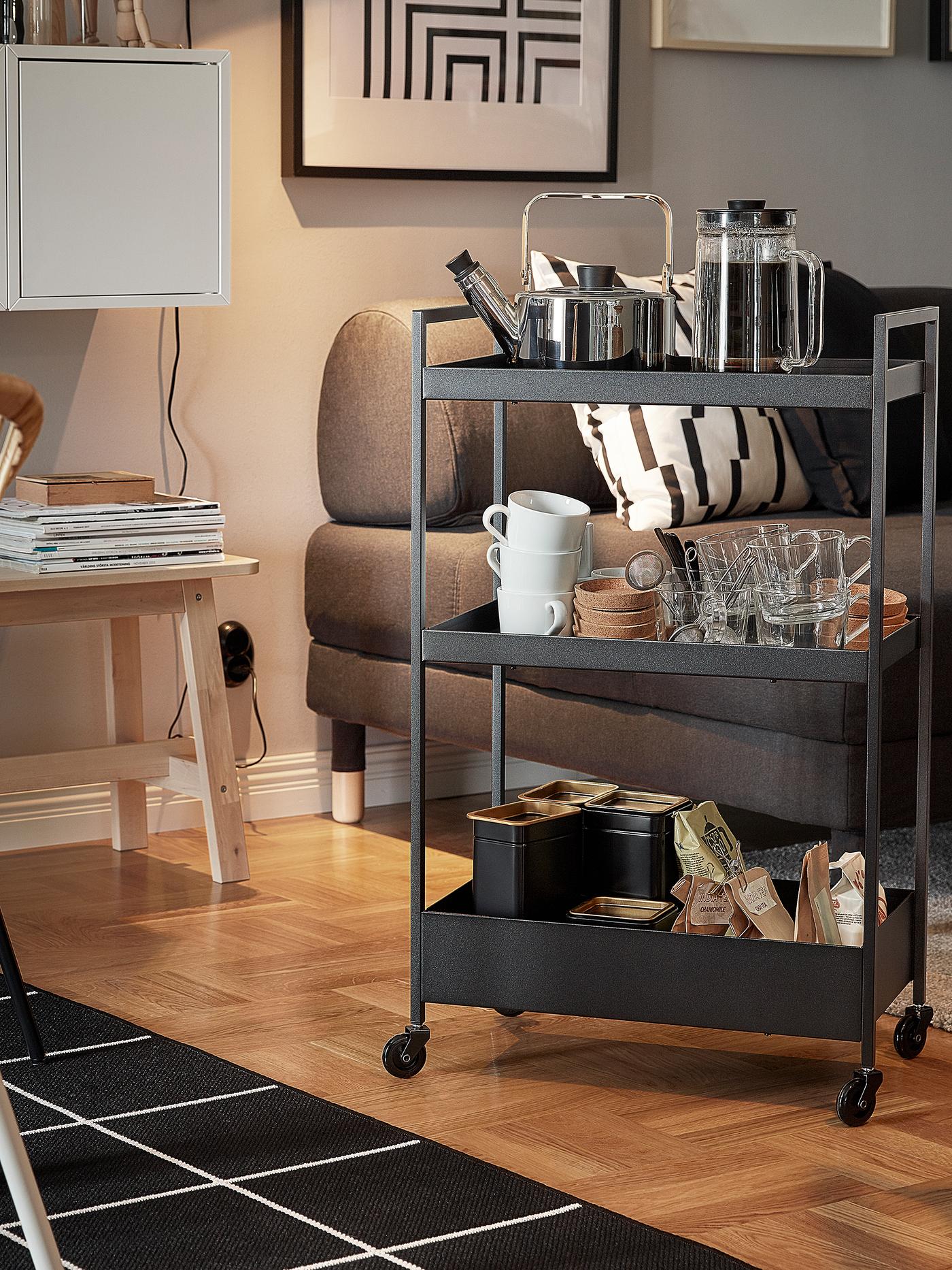A black NISSAFORS trolley with a kettle, mugs and tea bags on its shelves is standing next to a sofa and armchair.