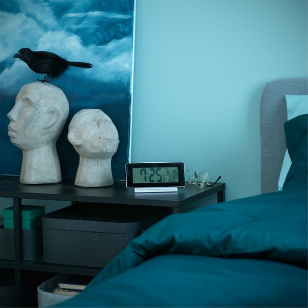 A black table with an alarm clock and decorative items is standing next to a bed with dark green bed textiles.