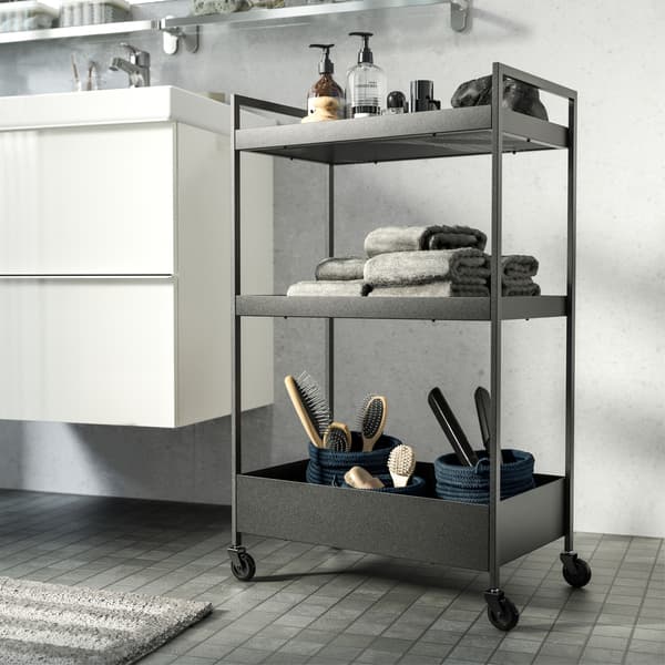A black trolley in a white/grey bathroom. It stores soap, towels and brushes and stands next to the wash basin.