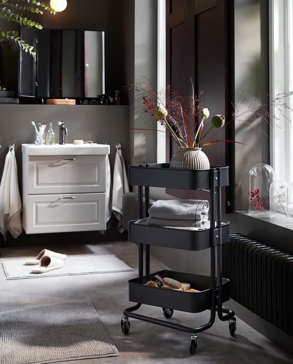 A black trolley on castors with a vase and towels on the shelves. Behind is a white wash-stand/wash-basin.