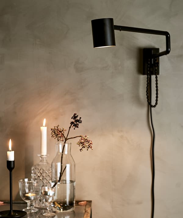 A black wall light on a painted grey wall, shining light on a glass counter with a mix of glassware, candles and tealights.