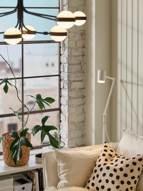 A black/white STORSLINGA chandelier in a room with a white sofa, a white floor/reading lamp and a pot plant in the window.