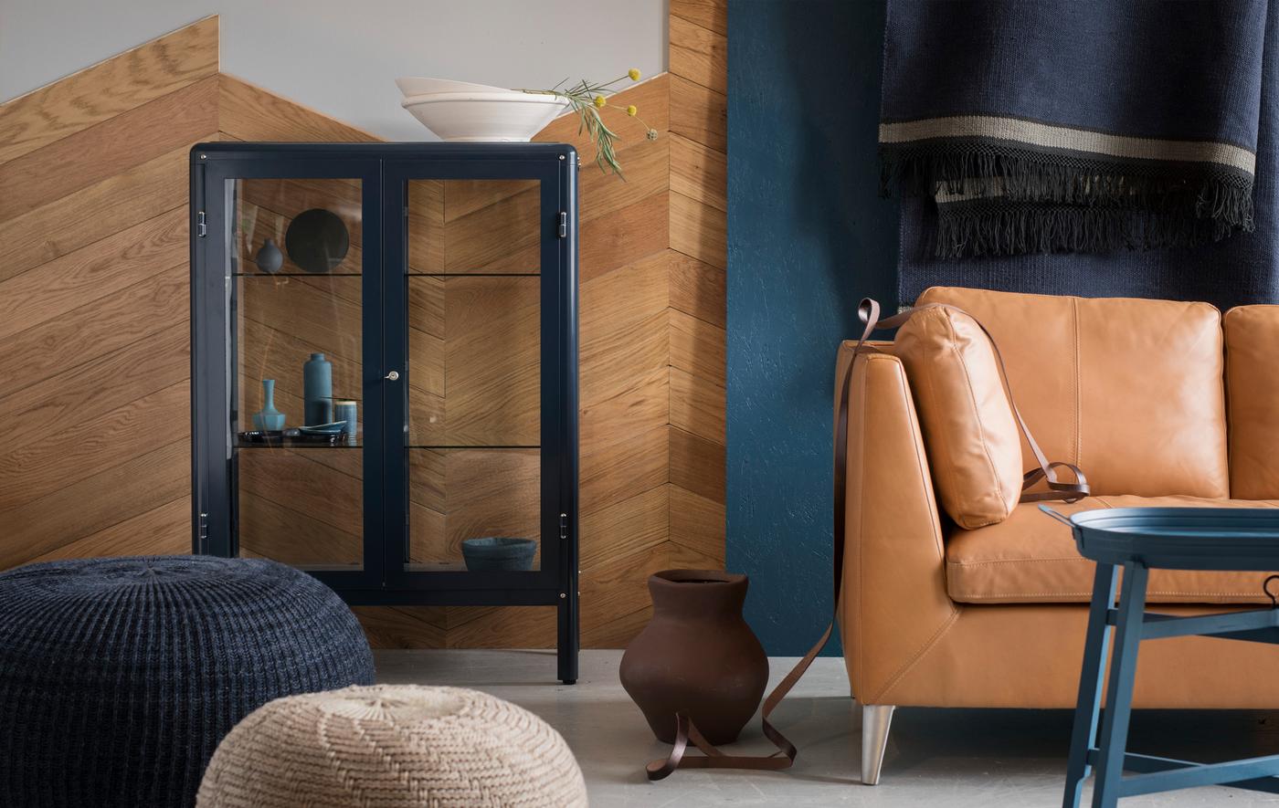 A blue and brown living area with a sofa, footstools and an IKEA FABRIKÖR glass-door cabinet in blue.