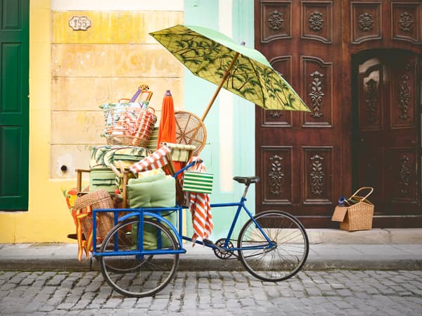 A blue bicycle standing on a cobblestone road, stacked with beach style furniture from the IKEA SOLBLEKT collection.
