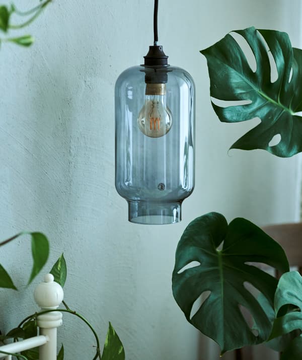 A blue glass pendant lampshade hanging beside a white bedframe, next to a MONSTERA plant.