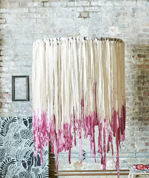 A boho mobile hangs by a brick wall. The hoop is decorated with tassel-like strips of white fabric, dyed pink at the ends.