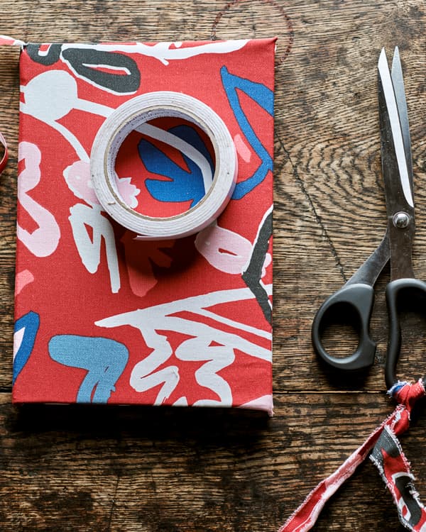 A book covered in red patterned fabric on a rustic wood table with a roll of sticky tape on top, next to a pair of scissors.
