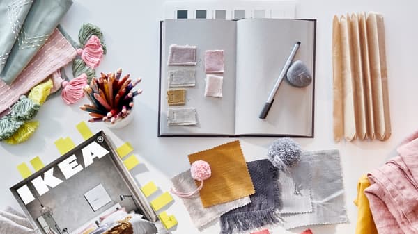 A bookmarked IKEA catalogue, an open notebook, a cup of coloured pencils and fabric samples in various colours.