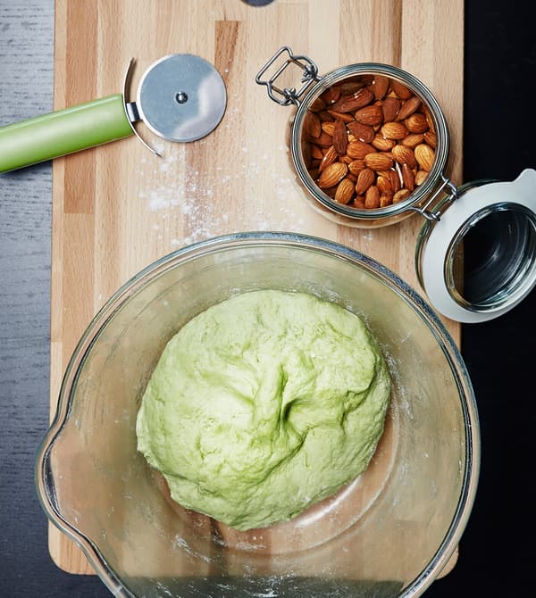 A bowl of green dough sits in a glass bowl on a wooden chopping board.