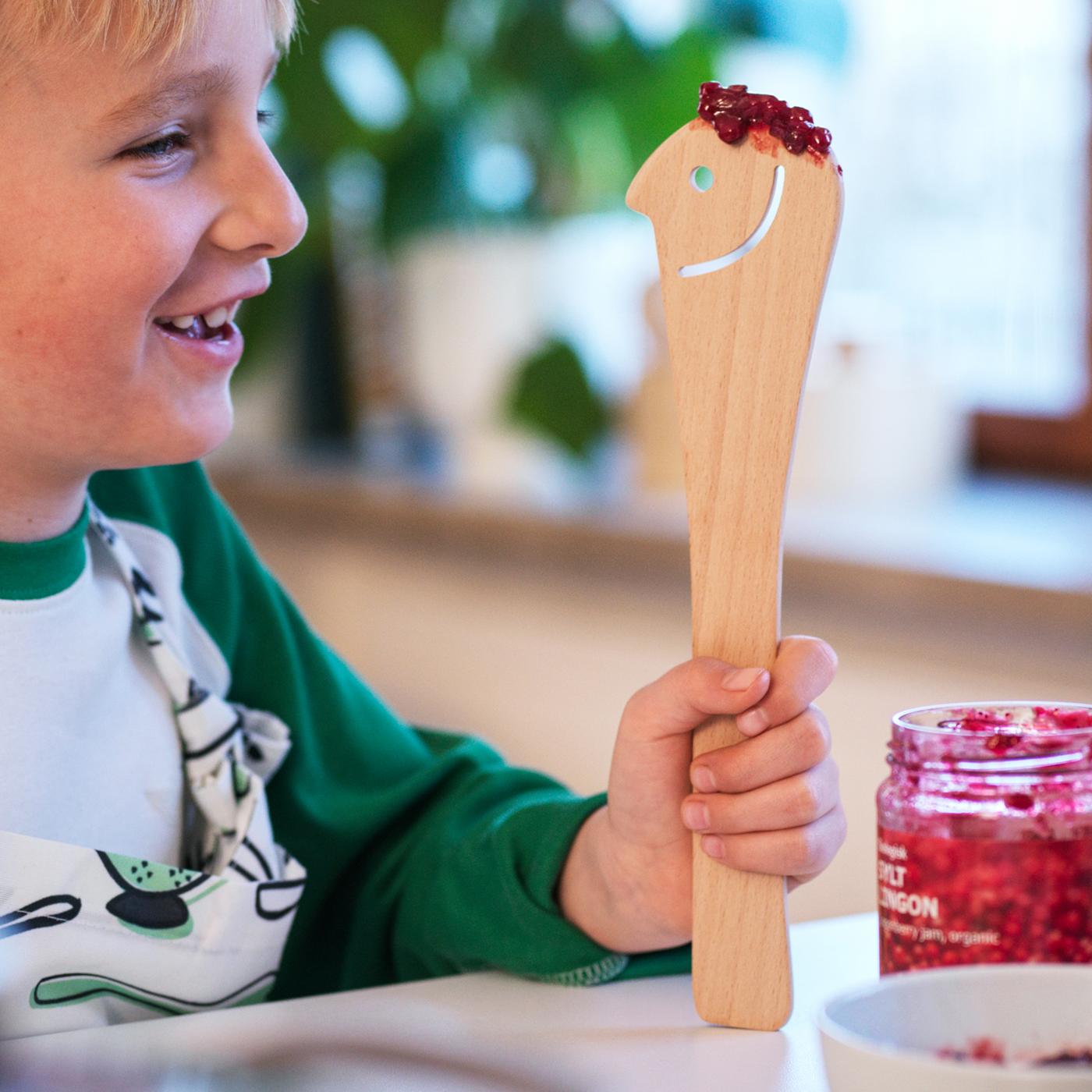 A boy holds a wooden UPPLIVAD spoon smeared with SYLT LINGON spread on a kitchen counter; the spoon has a smiley face cut-out.