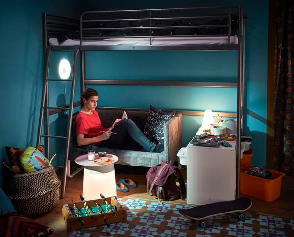 A boy sitting in a small sofa doing his homework. The sofa is placed under a loft bed.