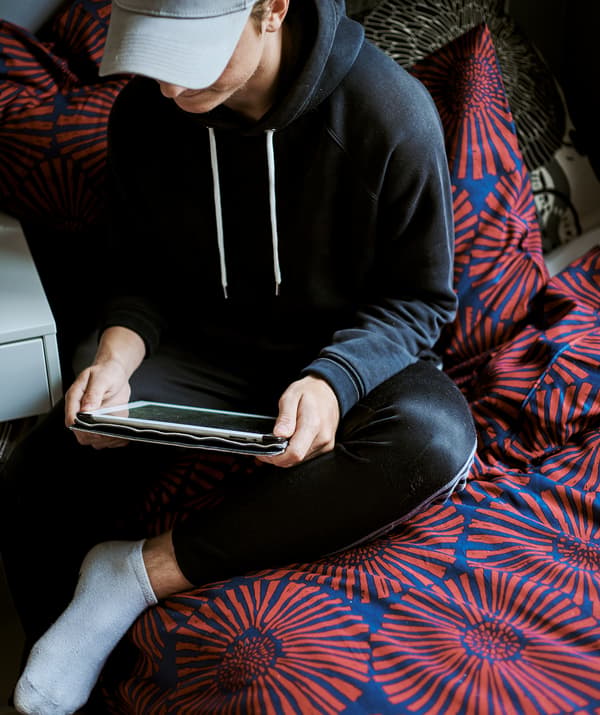 A boy wearing a baseball cap, playing on a tablet sits on a bed with a graphic printed red and blue duvet.
