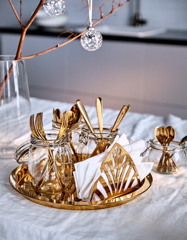 A brass-coloured tray carrying brass-coloured cutlery in jars and white napkins in a brass-coloured napkin holder.