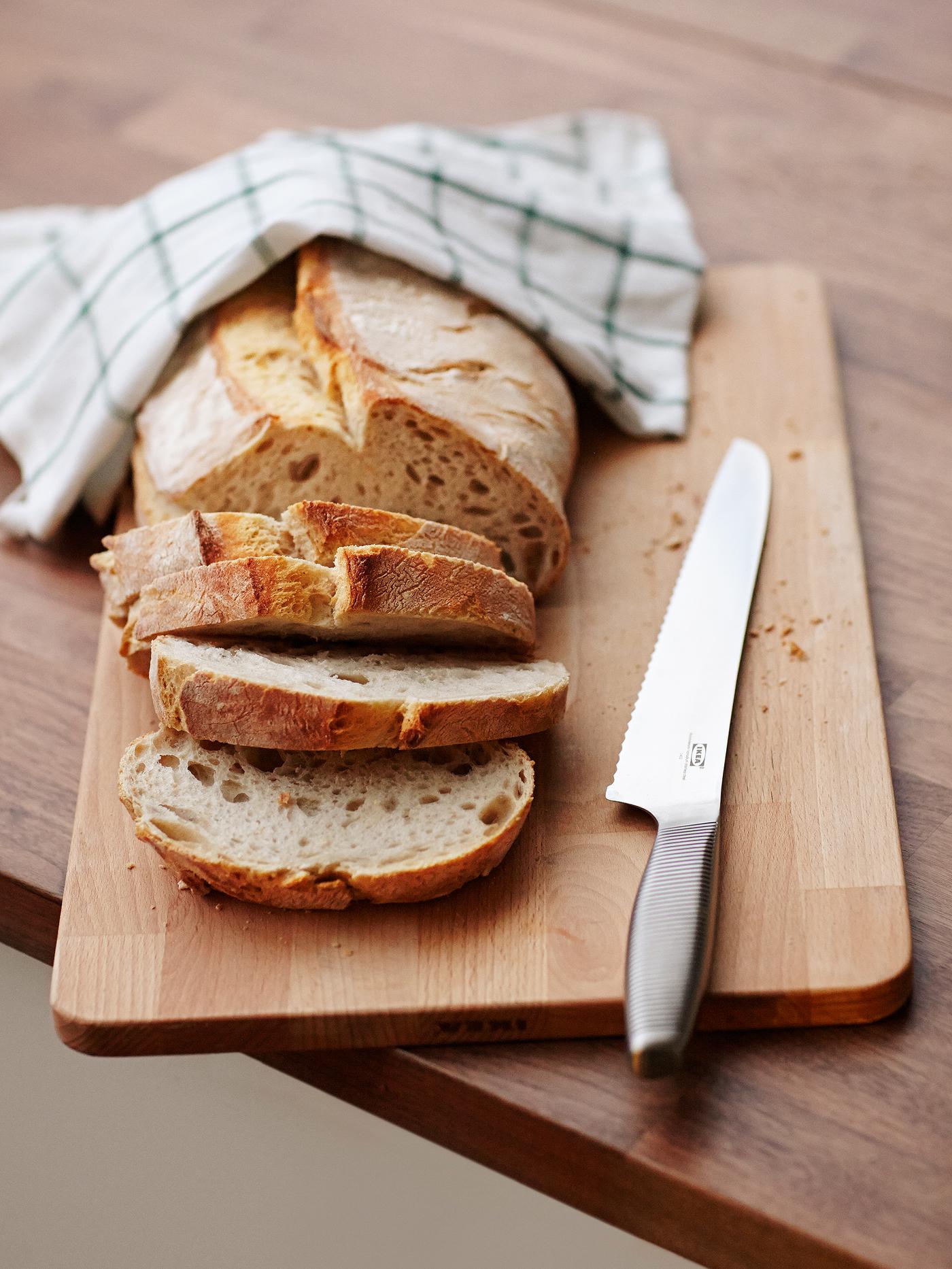 A bread knife on a wooden chopping board next to a loaf of bread where some slices have been cut and a tea towel.