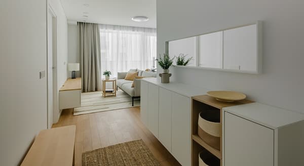 A bright and modern hallway leading into a cozy living room with floor-to-ceiling windows covered by sheer curtains. The space features minimalist white cabinetry, warm wooden floors, a wall mirror, decorative plants, and soft lighting that enhances the clean, Scandinavian-inspired design.