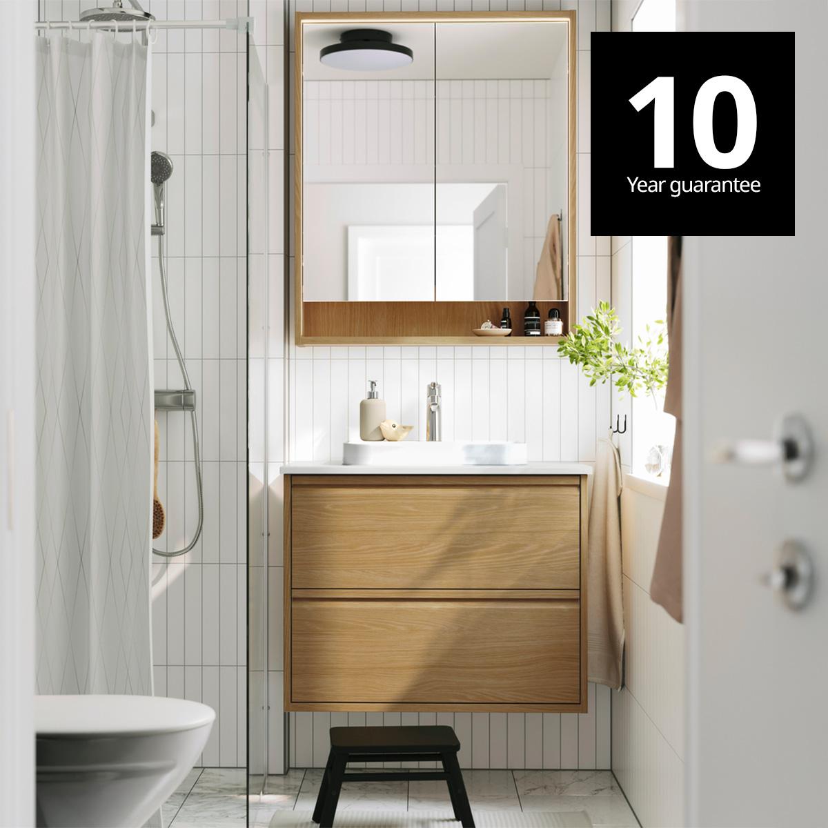 A bright bathroom has ÄNGSJÖN bathroom furniture in oak effect, including wall cabinets and a wash-stand with drawers. 10-year guarantee logo on top right corner.