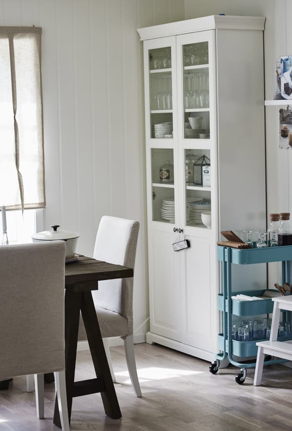 A bright blue IKEA trolley parked next to a white kitchen cabinet with glass doors displaying glassware and tableware.