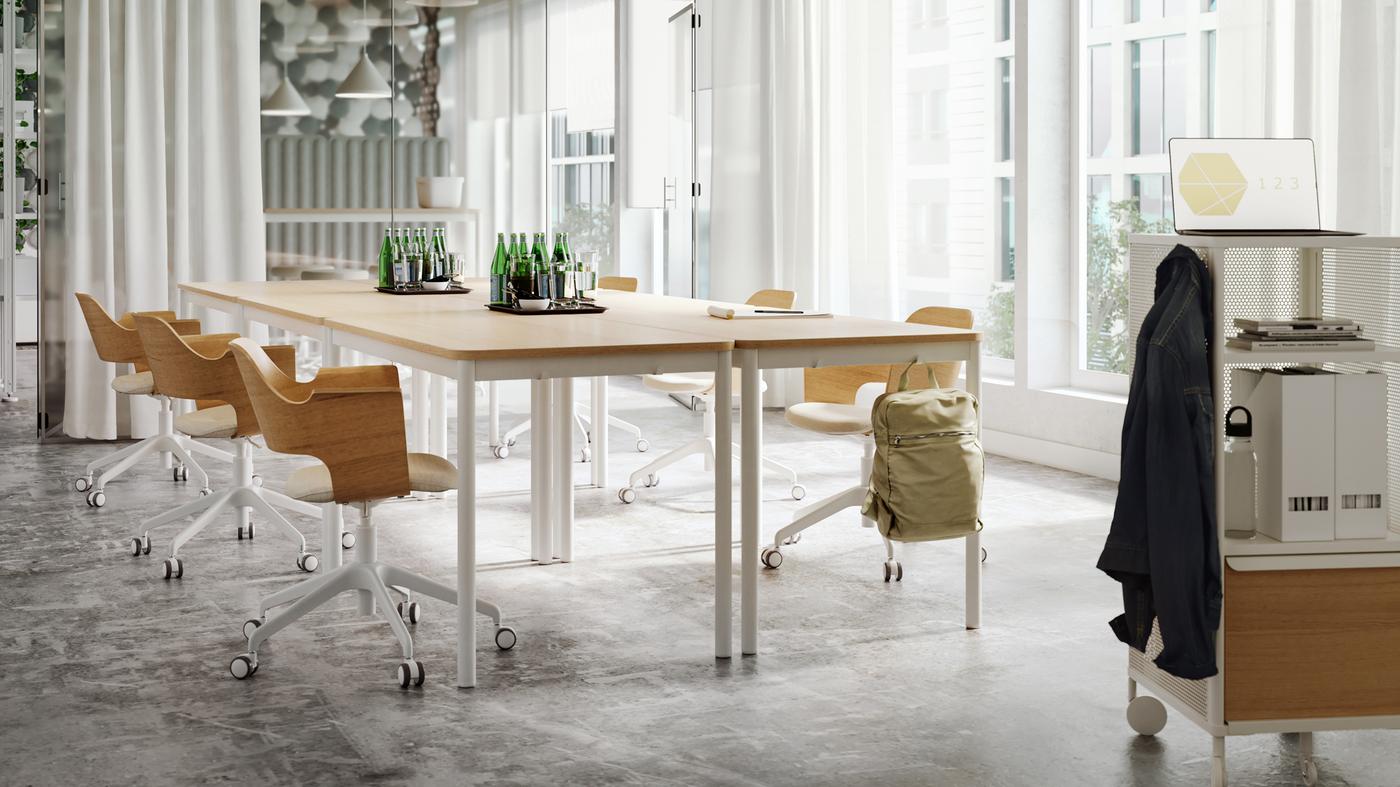 A bright meeting room with tables and chairs in light wood and white tones, sheer white curtains and a glass wall.