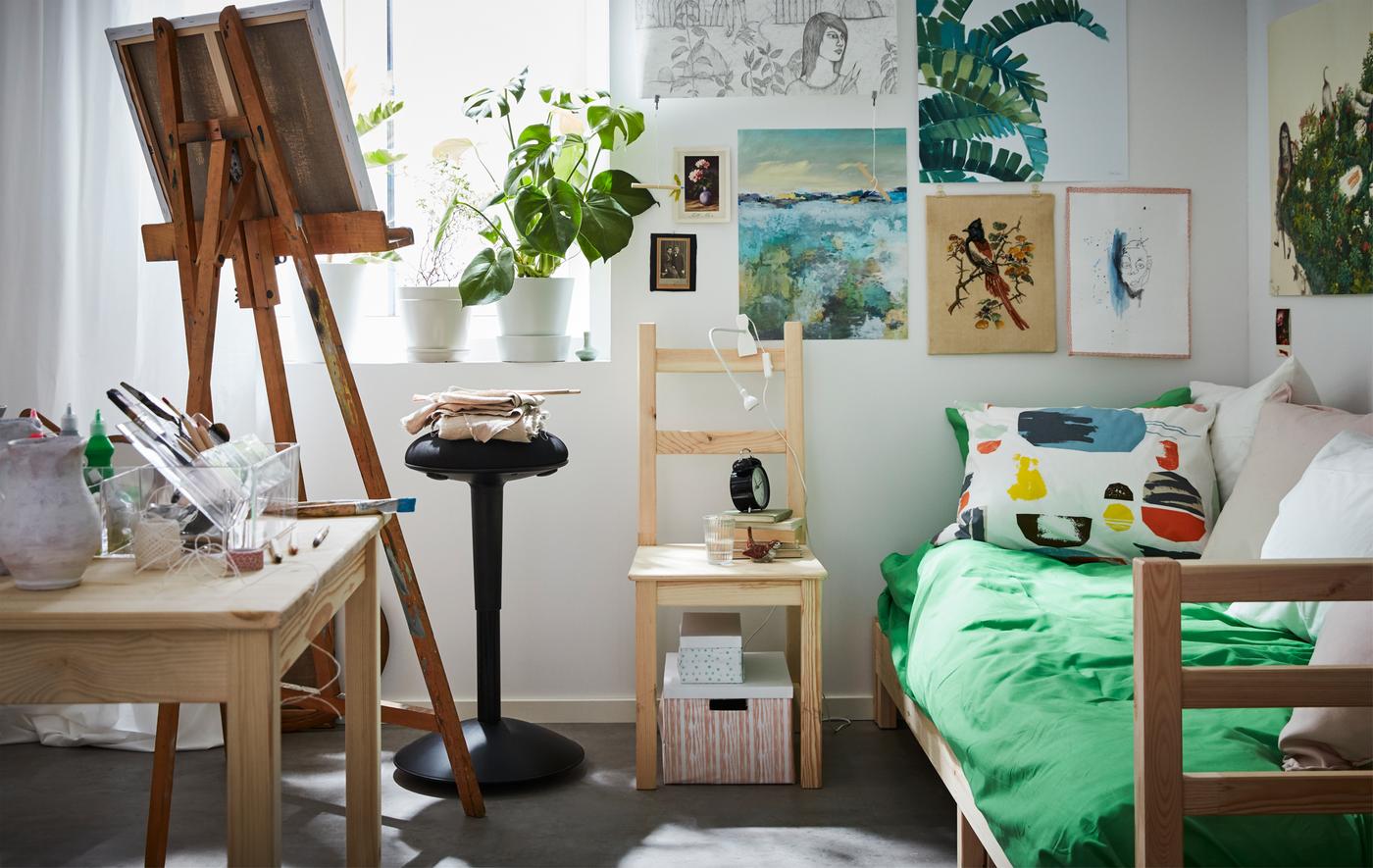 A bright, white dorm room where an artist lives. There are colourful posters on the walls, a bed, and an easel.