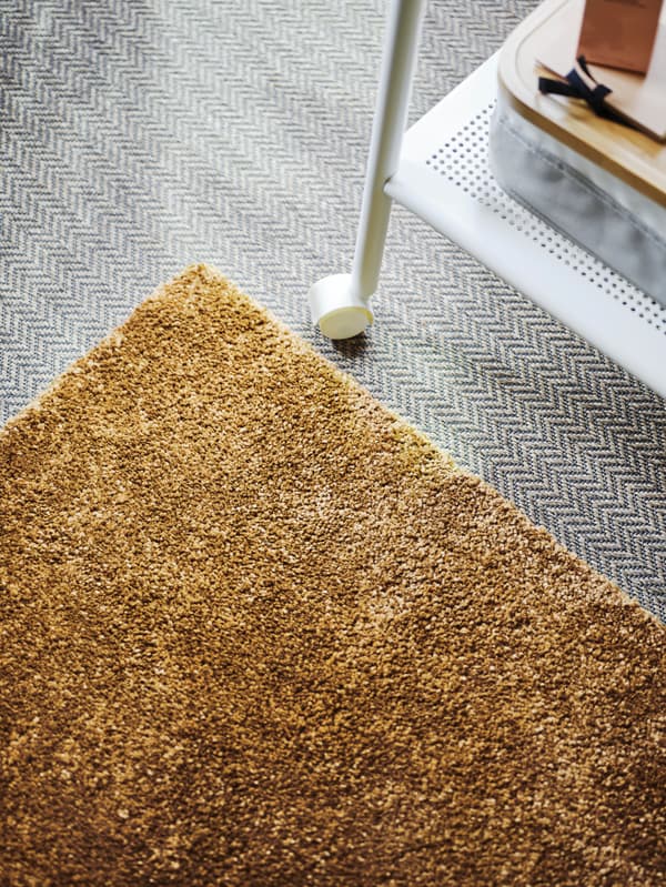 A brown-yellow STOENSE rug is placed on a grey floor, with a white metal rolling table alongside it.