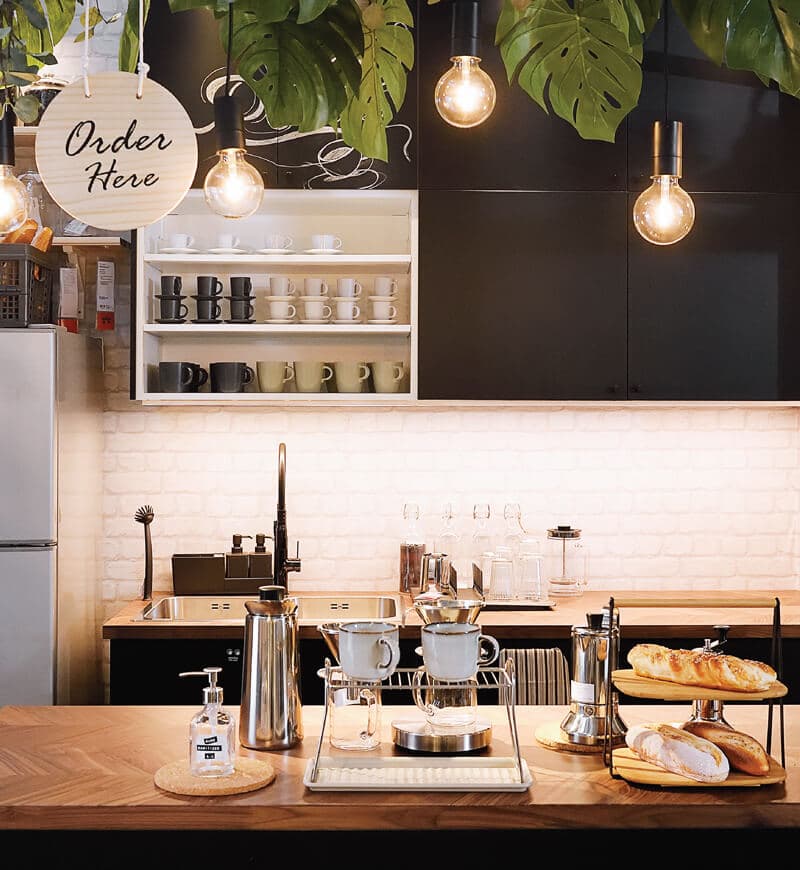 A café ordering area featuring two wooden countertops with coffee making accessories and plants at the top of the ceiling.