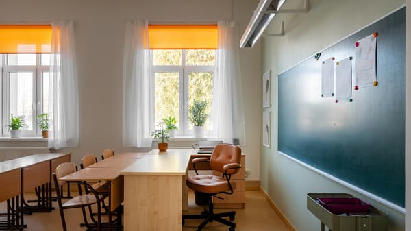 A calm and functional study corner within an after-school space, featuring wooden desks, chairs, a teacher’s chair, and a large chalkboard wall. Soft natural light, orange blinds, and light-toned furnishings create a warm and welcoming atmosphere.