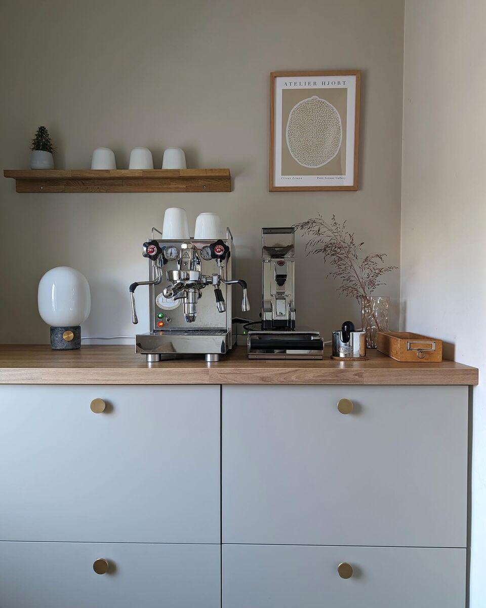 A calm, minimalist coffee corner with an espresso machine and coffee grinder on a light grey sideboard with a wooden top, white cups on a wall shelf, a small table lamp, dried flowers, and a framed poster against a neutral wall.