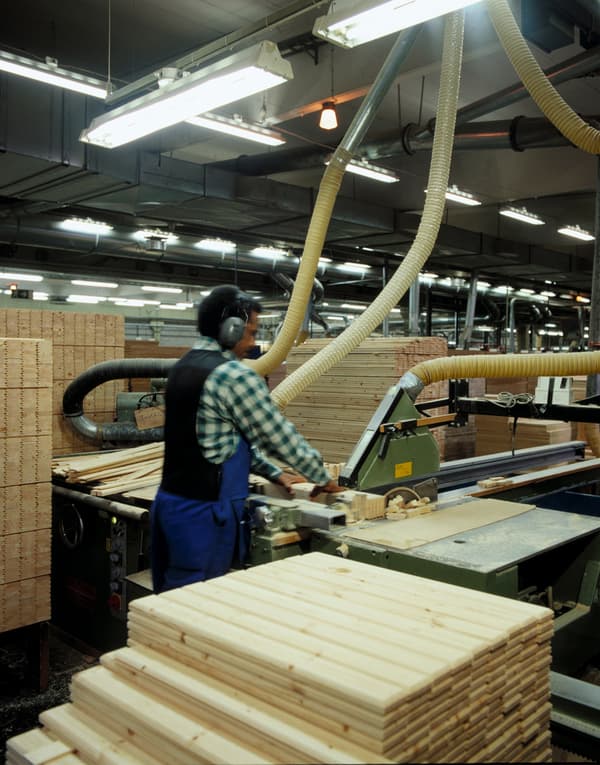 A carpenter wearing ear protection has piles of wooden boards around him while sawing wood with a machine in a factory.