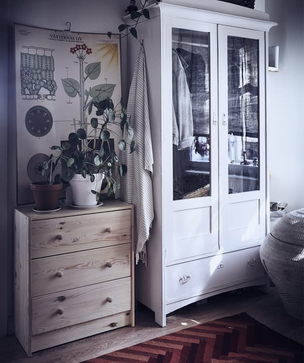A chest of drawers and wardrobe in a hallway, with plants and a wall hanging above the chest.
