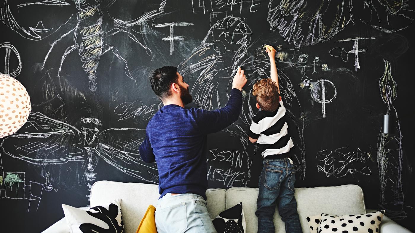 A child and adult kneel and stand on a sofa drawing pictures on a blackboard wall.