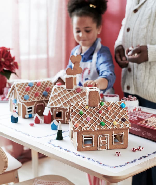 A child and an adult standing by a table on which is placed several finished gingerbread houses neatly decorated with icing.