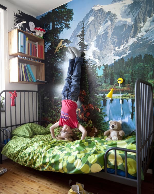 Á child does a headstand on a bed and rests his feet against a wall with a mountain scape.