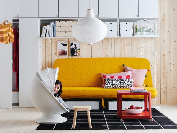 A child hiding in an egg chair in front of a yellow NYHAMN sofa. A red TINGBY side table on castors over a checkered rug.