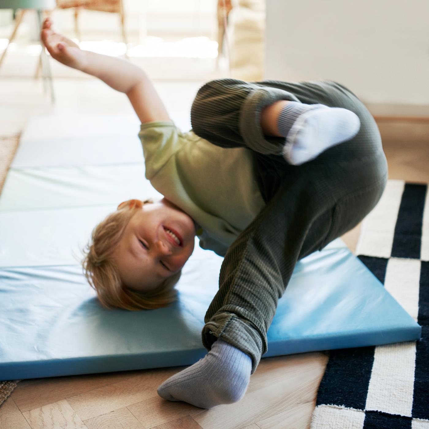 A child in dark green pants and a light green top does a tumble on a blue PLUFSIG folding gym mat.