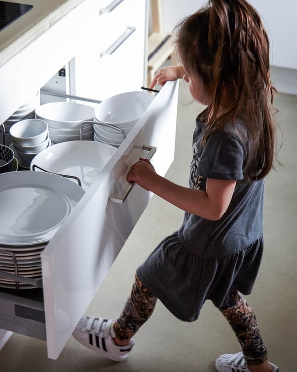 A child opening a large white kitchen drawer to reveal stacks of white dishware.