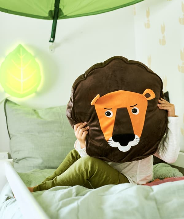 A child sits on a bed under a leaf-shaped canopy holding a cushion in the shape of a lion in front of their face.