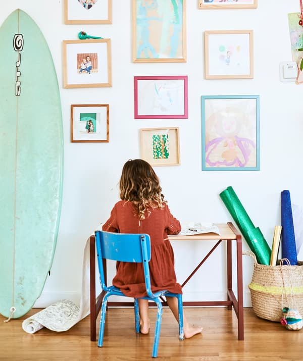 A child sits on a blue chair at a small desk, against a wall of children’s paintings and family photographs next to a surfboard.