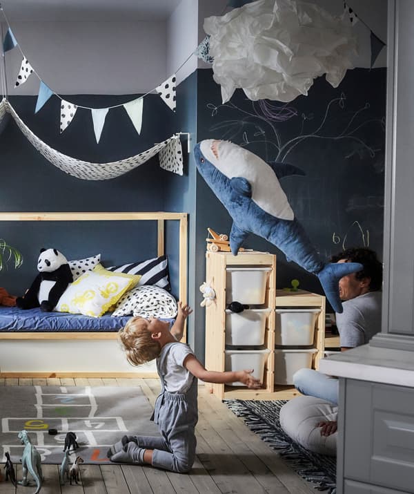 A child throwing a soft toy shark in the air, with storage boxes and a fabric canopy behind.