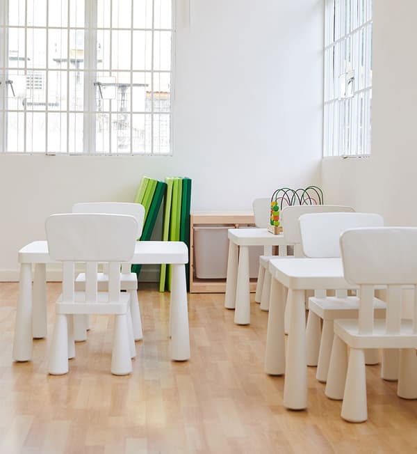 A children classroom with small white tables and chairs placed on a wooden tables.