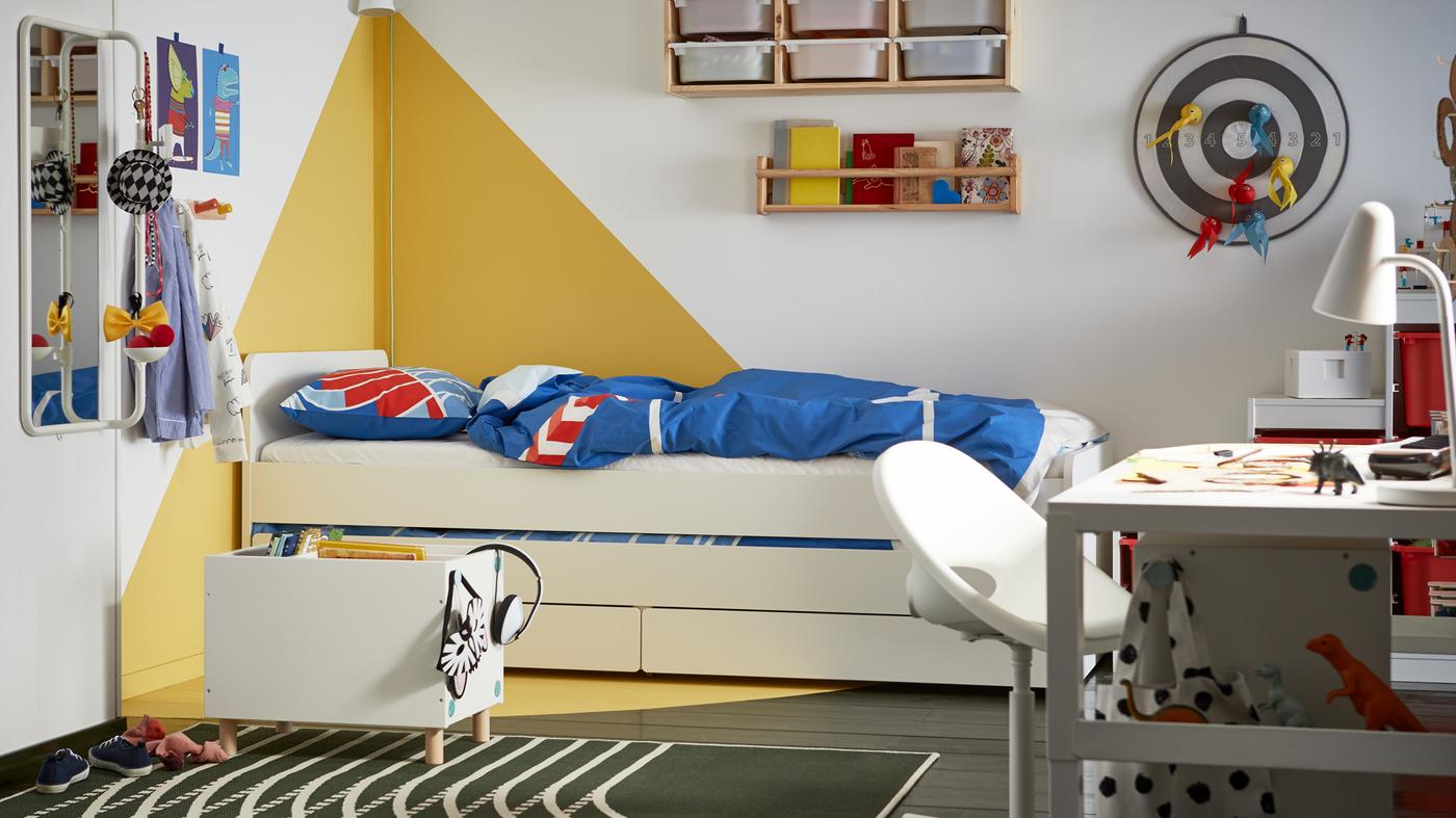A children's bedroom with graphic white and yellow walls, a white bed with storage underneath, blue and red bed linen.