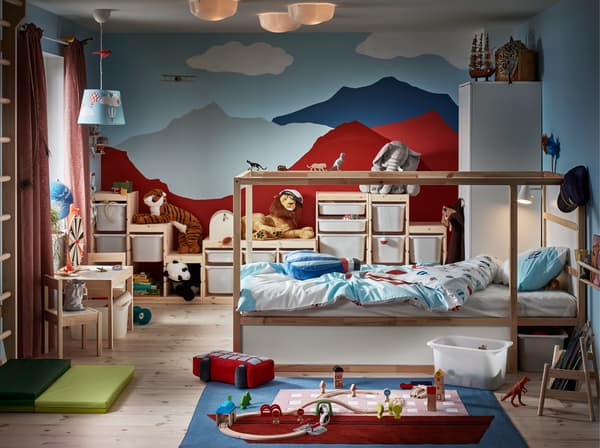 A children's room with a bed in pine, a wall that's painted like mountains, a rug with a boat pattern and lots of soft toys.