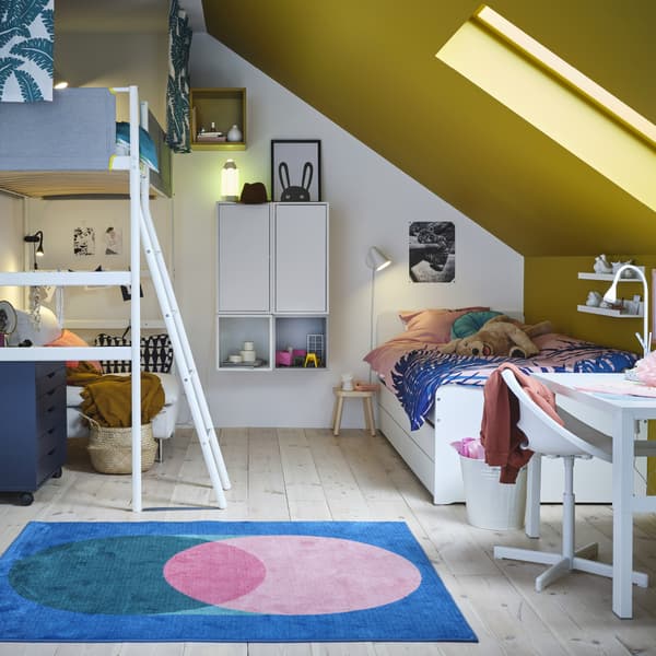A children’s room with a loft bed, a white bed frame, wall-mounted units and cabinets, a white desk and a pink/green rug.