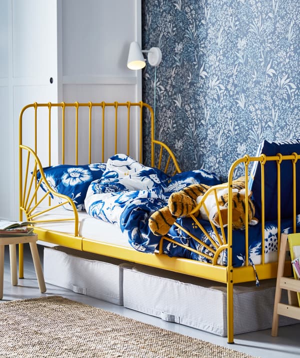 A child’s bed with yellow frame and blue floral bedding, with boxes underneath and a wall lamp above.
