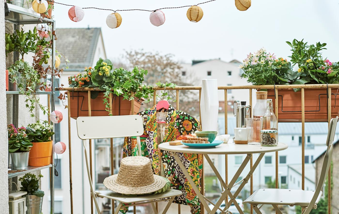 A city balcony with flower boxes, a shelf unit with plants, string lights and a bistro-style dining set, with snacks set out.